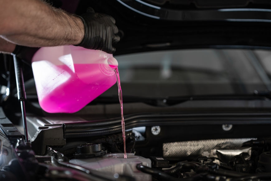 Pessoa com luva preta despejando l&iacute;quido rosa de radiador no compartimento do motor de um carro aberto.